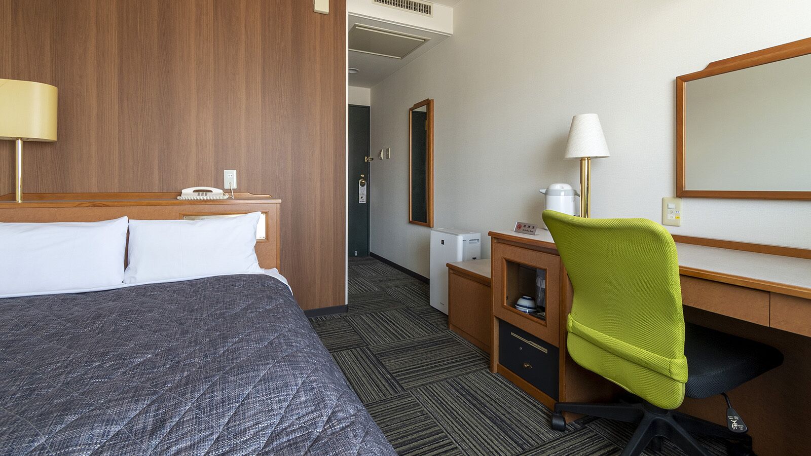 Hotellihuone, jossa on sänky, työpöytä, tuoli, peili, lamppu ja pieni keittokomero.