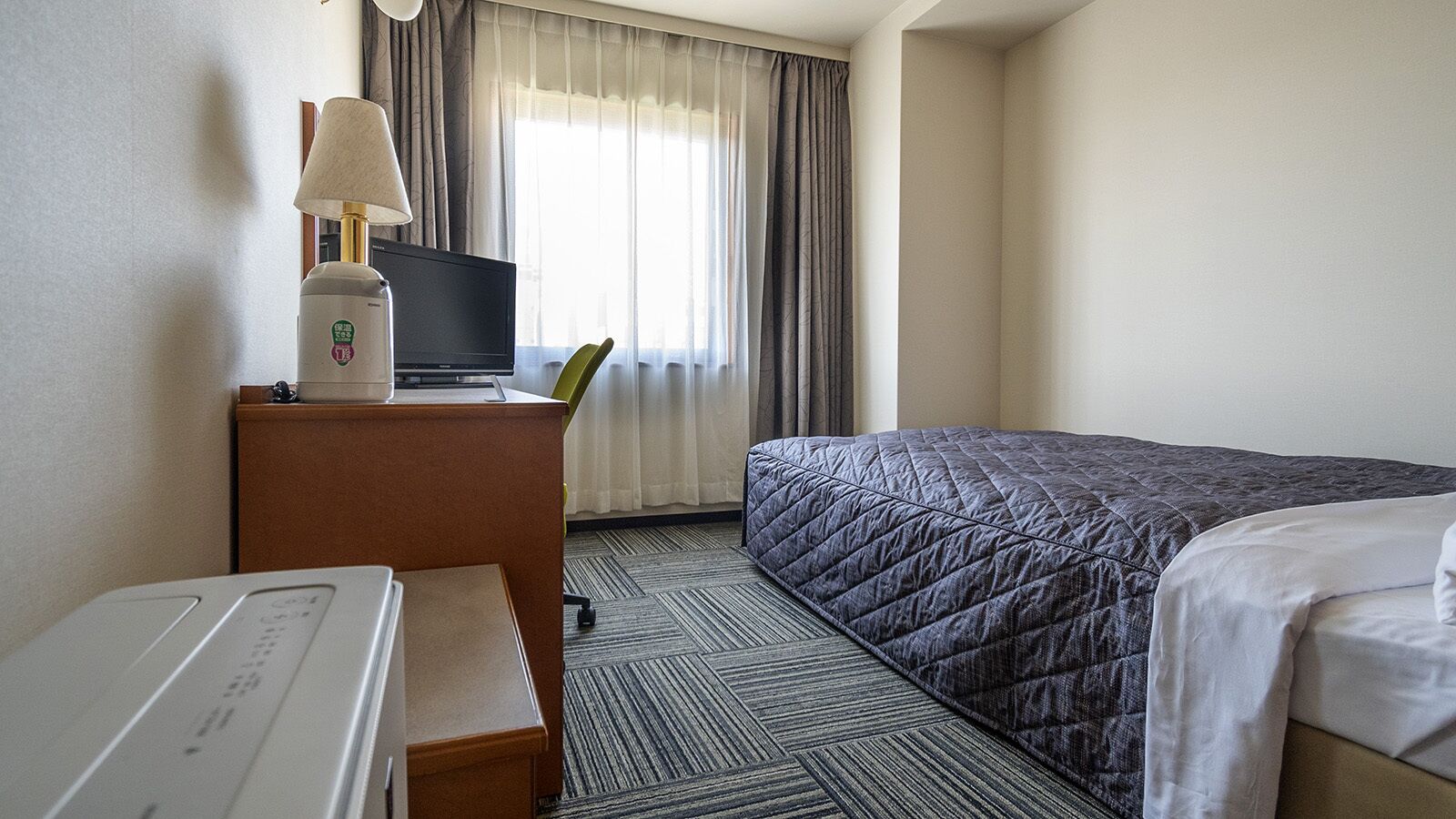 Hotellihuone, jossa on sänky, työpöytä lampun kanssa, televisio ja ikkuna, jossa on verhot.