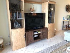 Uma sala de estar com uma TV sobre um armário de madeira, uma mesinha com uma cadeira e um objeto decorativo na parede.