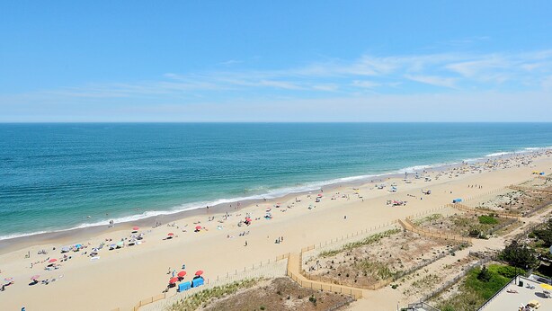 A beach with a fence, umbrellas, and people. - XPLORIE ACTIVITIES & LINENS INCLUDED*!  PLAZA - 2BR/open den. Indoor/outdoor pools & Tiki Bar. (Ocean City)