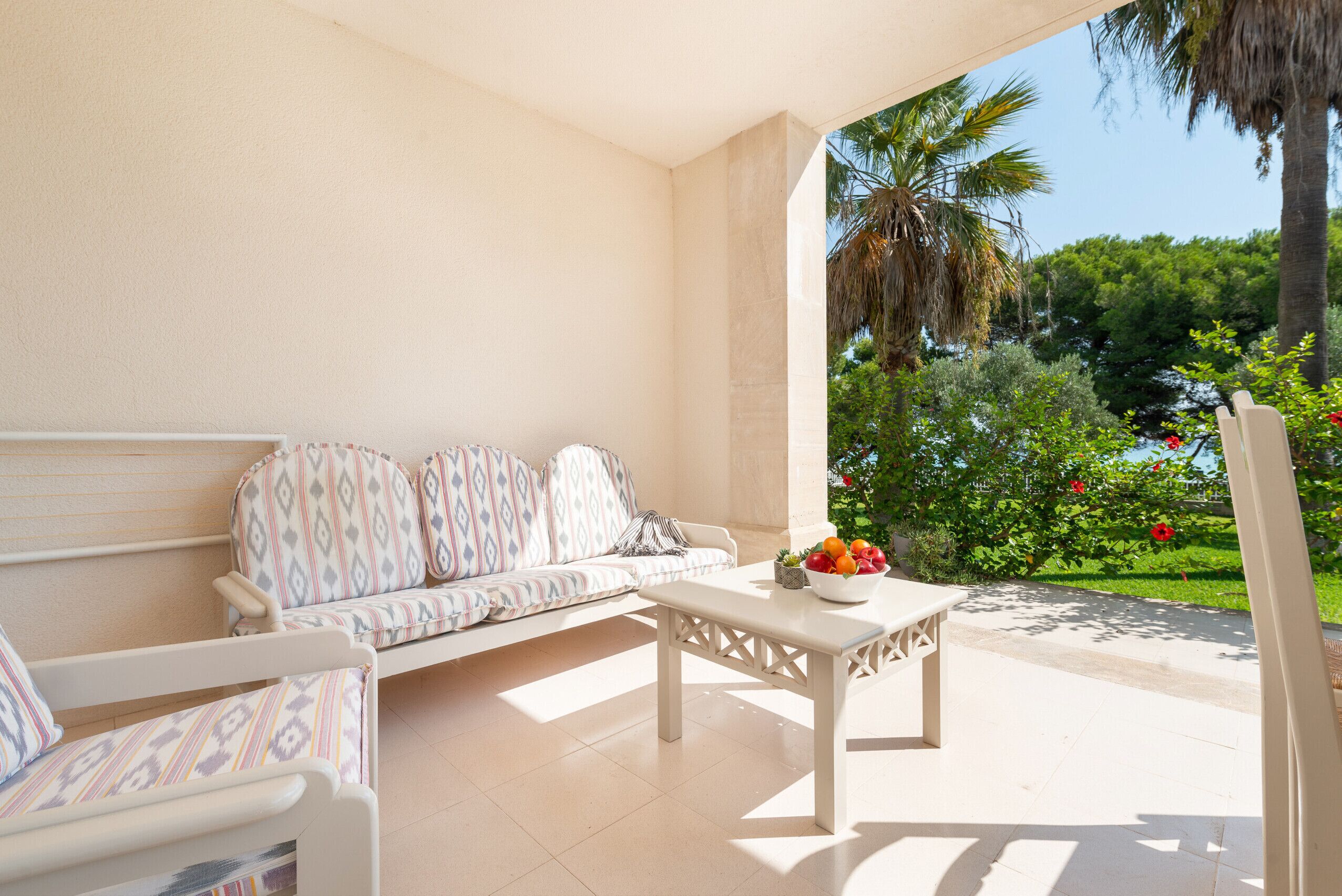 A sunlit patio with a white sofa, a small table with fruit, and a view of lush greenery and a palm tree.