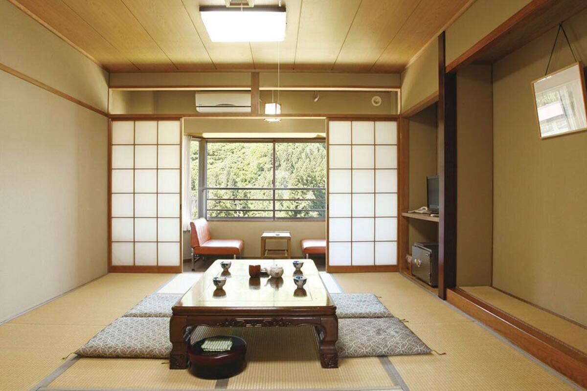 Una habitación japonesa tradicional con suelo de tatami, una mesa baja con juego de té, una puerta corredera y vistas a la vegetación exterior.