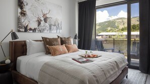 A bedroom with a large bed, a painting of elk, a view of the outdoors, and a balcony with chairs. - Outpost: One Town Hill 506 - Base of Snow King Mtn (Jackson)