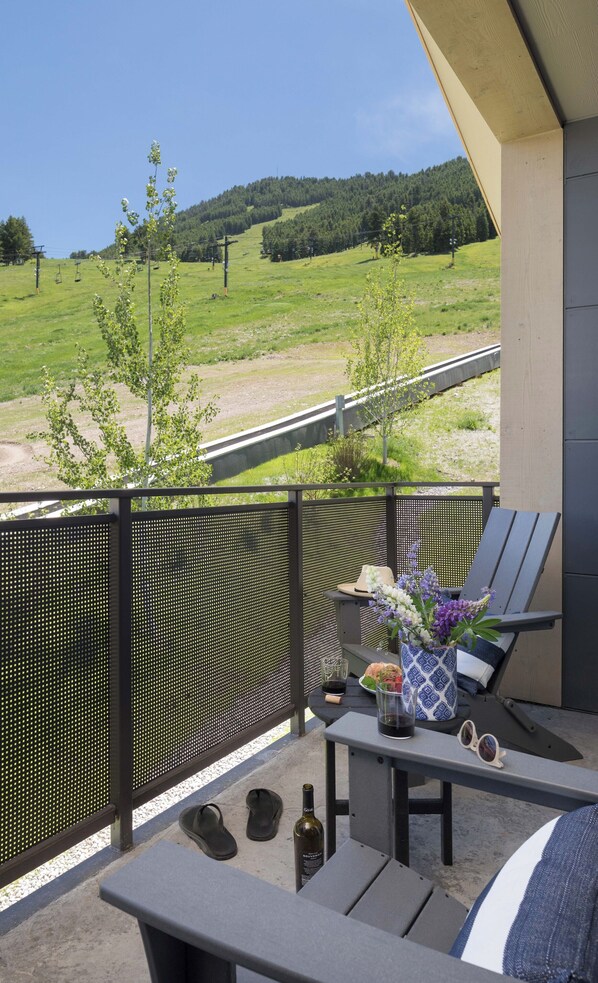 A balcony with a view of a green field and a forest. There is a table with a bottle, glasses, and a vase with flowers.