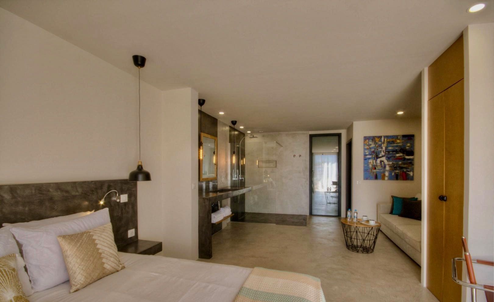 A modern hotel room with a large bed, a bathroom with a glass shower, and a living area with a sofa and a painting.