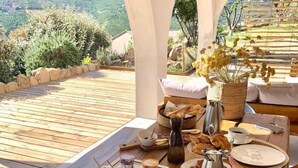 Ein Frühstückstisch auf einer Holzterrasse mit schöner Aussicht, mit Früchten, Gebäck, Kaffee und einer Blumenvase.