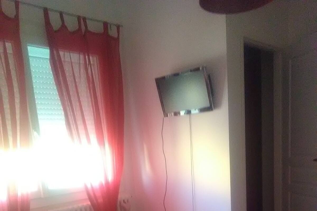 A hotel room with a bed, red curtains, a TV mounted on the wall, and a window with blinds.