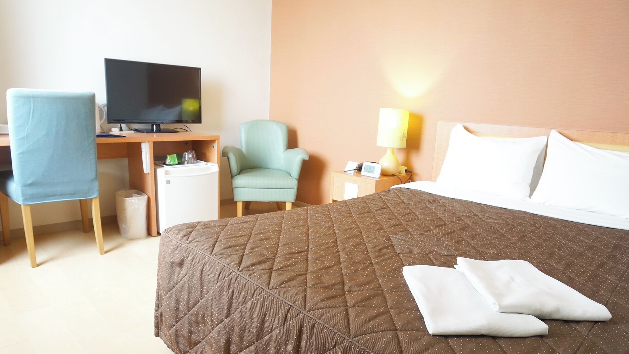 A hotel room with a bed, a desk with a TV, a chair, a nightstand with a lamp, and a small refrigerator.
