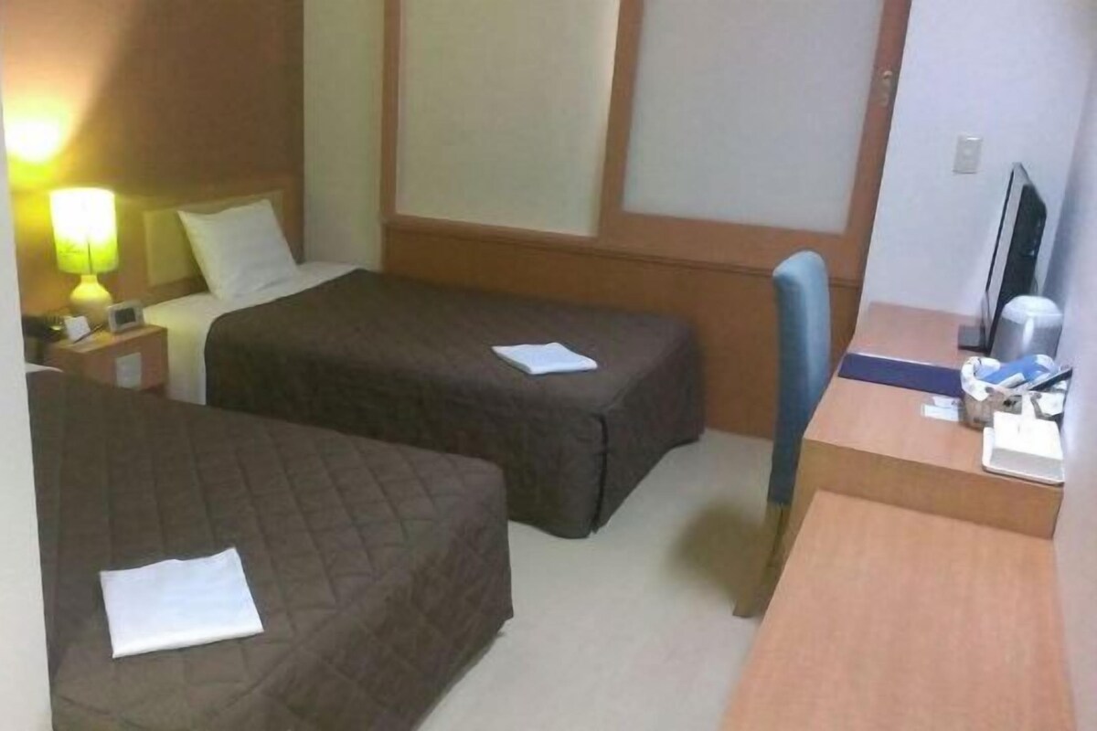 A hotel room with two single beds, a desk with a chair, a lamp, and a television.