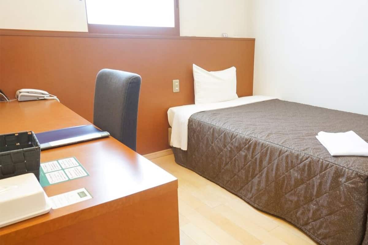 Quarto de hotel com uma escrivaninha de madeira, uma cadeira, uma cama com colcha acolchoada e uma janela.