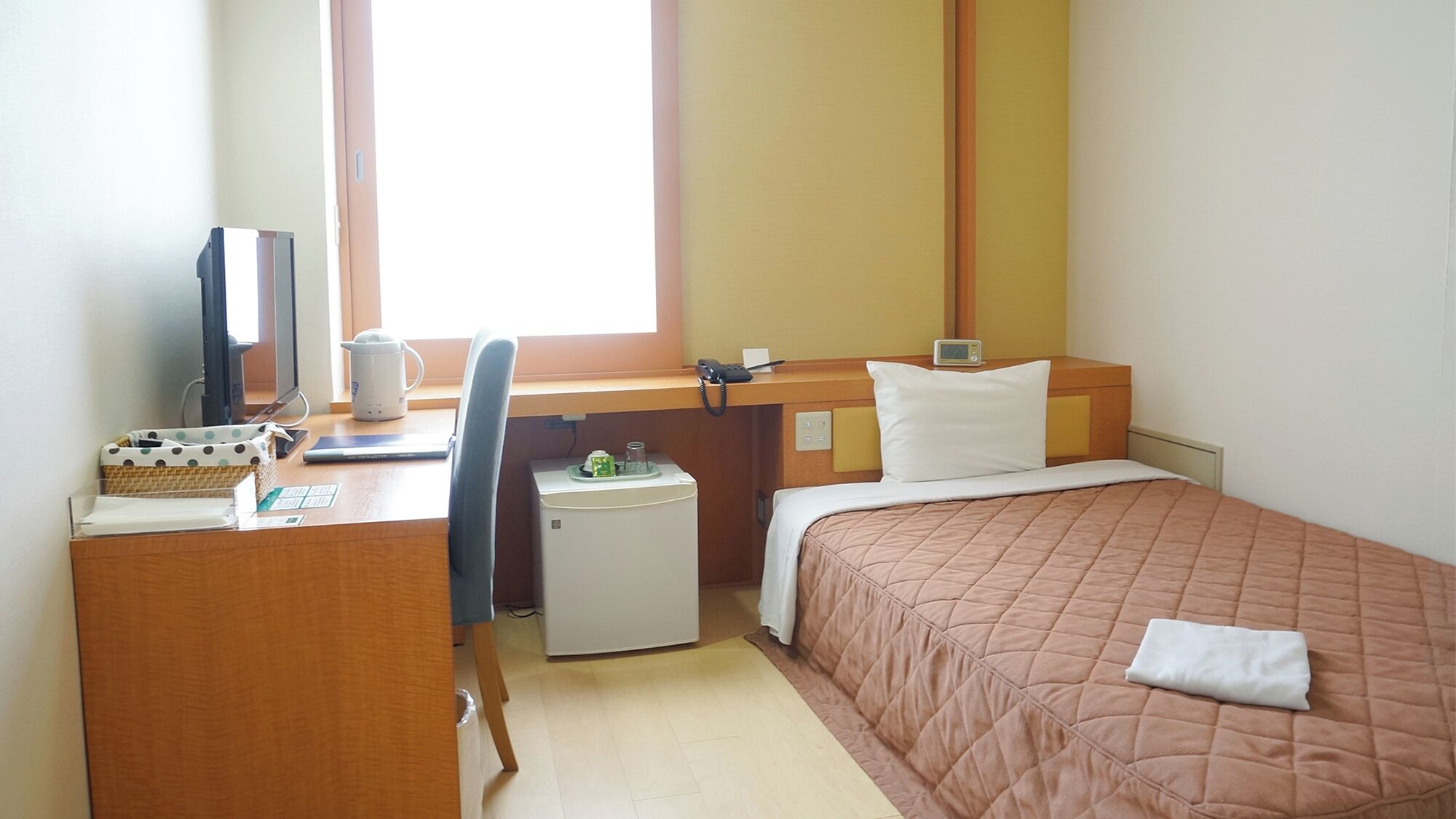 A hotel room with a desk, a bed, a small refrigerator, a chair, and a window.