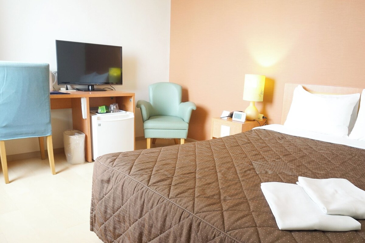 Habitación de hotel con cama, escritorio con televisor, silla, mesita de noche con lámpara y refrigerador pequeño.