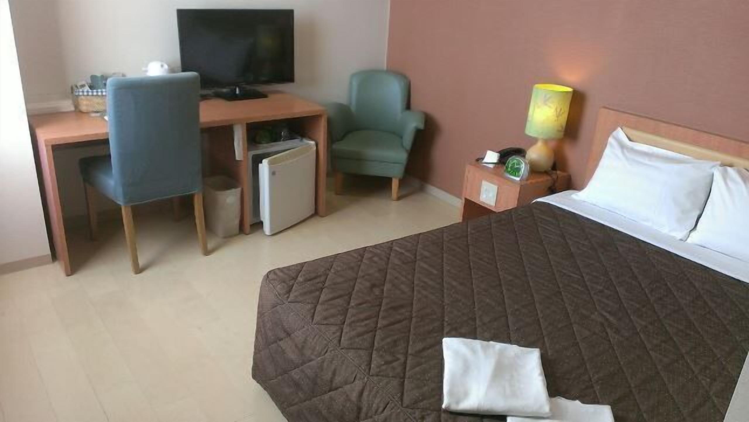 A hotel room with a bed, a desk with a computer, a chair, a lamp, and a nightstand with a clock.