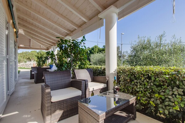 A covered outdoor seating area with wicker furniture and a glass table, surrounded by greenery and a clear sky.