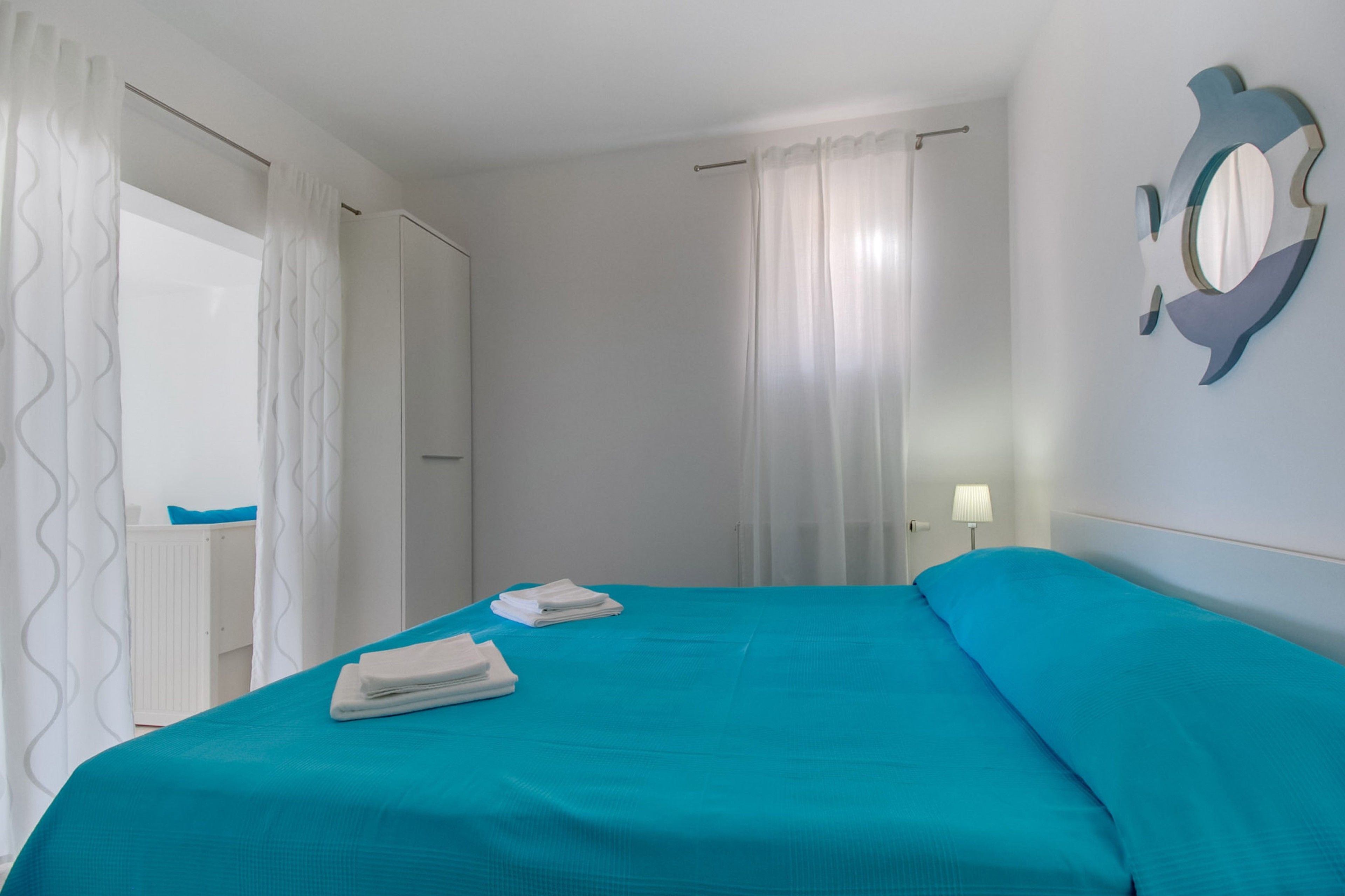 Une chambre d’hôtel avec un lit, des rideaux blancs, une lampe fixée au mur et une fenêtre également dotée de rideaux blancs.