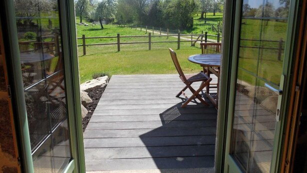 Eine Holzterrasse mit Tisch und Stühlen, ein eingezäunter Garten und freie Sicht auf die umliegende Landschaft.
