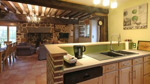 Eine geräumige Küche mit Kamin aus Stein, Holzbalken und einer großen Kochinsel mit Spüle und Geschirrspüler.
