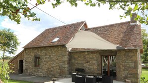 A stone house with a tiled roof, a covered patio with outdoor furniture, and a grassy yard.