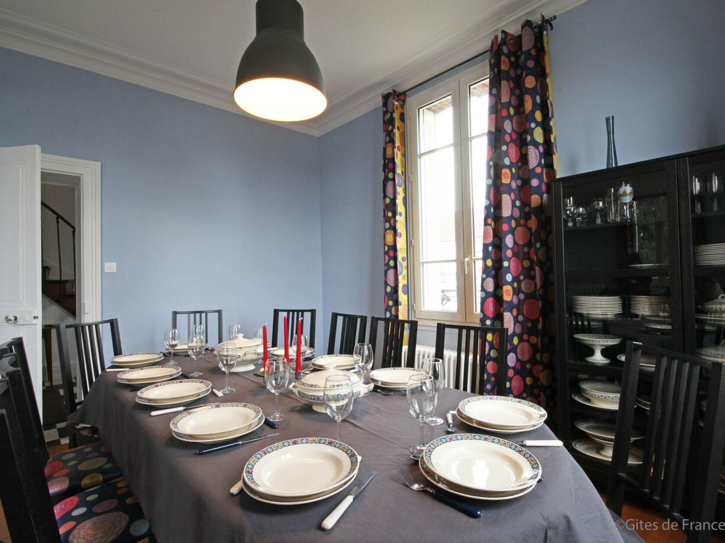 Ein Esszimmer mit Tisch für sechs Personen, Stühlen, einer Kandelaber, einem Fenster mit Vorhängen und einem Serverschrank mit Geschirr.