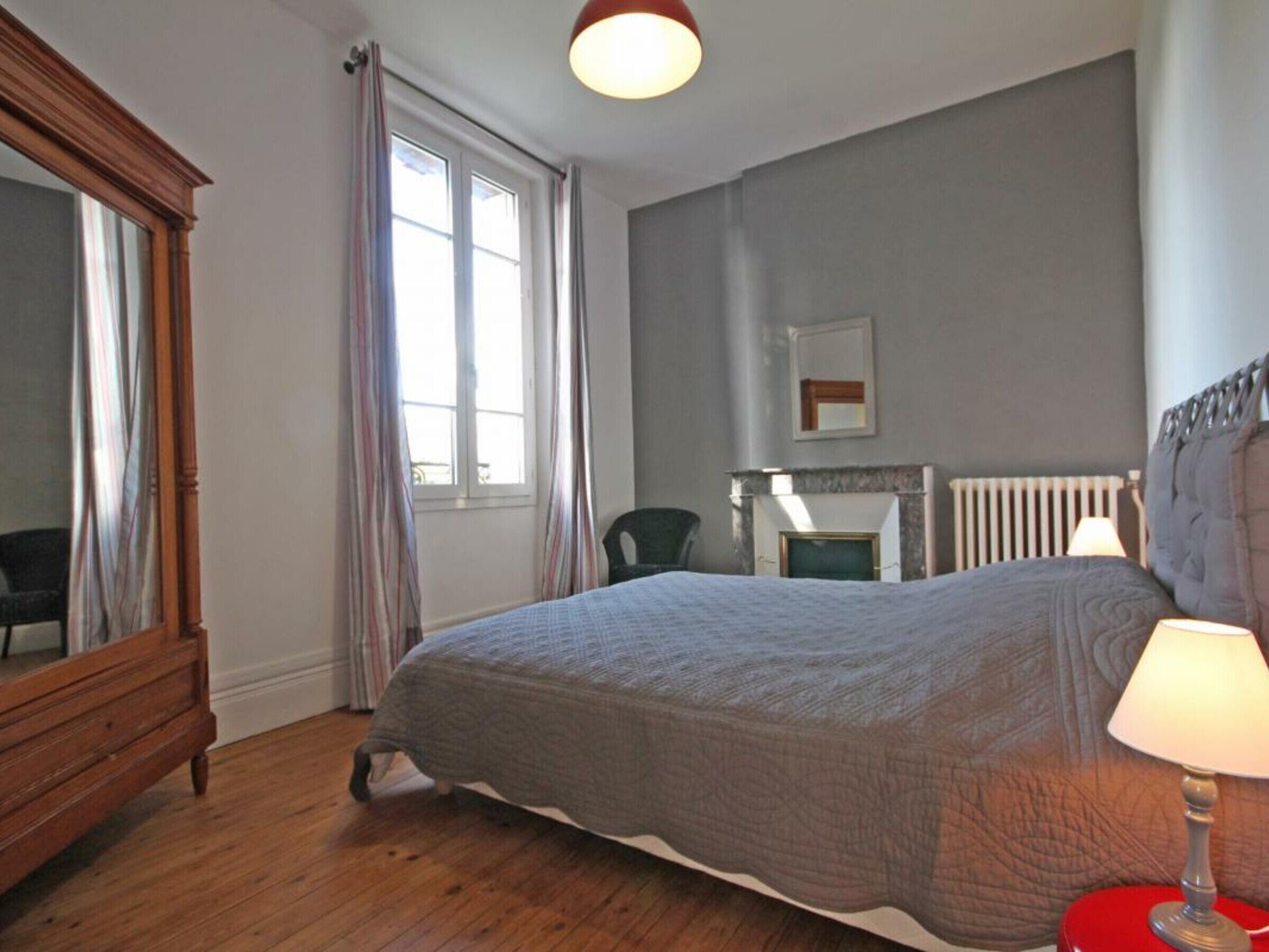 Ein Schlafzimmer mit Bett, Sessel, Kommode, Kamin und einem Fenster mit Vorhängen.