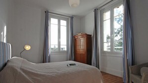 A bedroom with a bed, a desk with a lamp, a wooden wardrobe, and a chair.