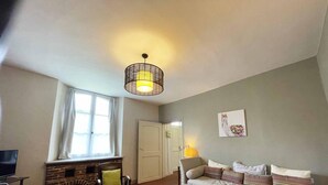 Een gezellige woonkamer met een bank, fauteuil en een houten bijzettafeltje. In de hoek is een open haard te zien, en een raam met gordijnen zorgt voor natuurlijk licht.