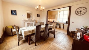 Een eethoek met houten vloer, rieten stoelen, een tafel gedekt voor vier personen, een kroonluchter en een keuken in de achtergrond.