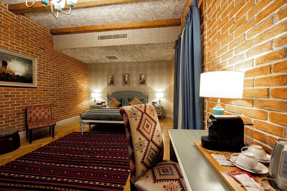 A hotel room with a brick wall, a bed, a chair, a table with a lamp, and a coffee maker.