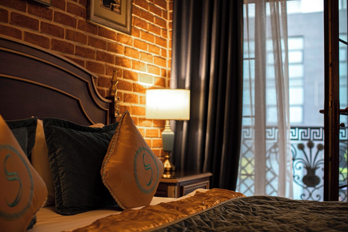 A hotel room with a brick wall, a bed with multiple pillows, a nightstand with a lamp, and a view of a balcony with a wrought iron railing.