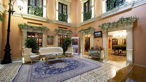 Un hall imponente con un tapiz grande y ornamentado, asientos lujosos y un candelabro.