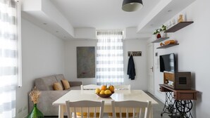 Een moderne woonkamer met een witte eettafel, gele stoelen, een grijze bank, een groene vaas en een televisie op een houten dressoir.