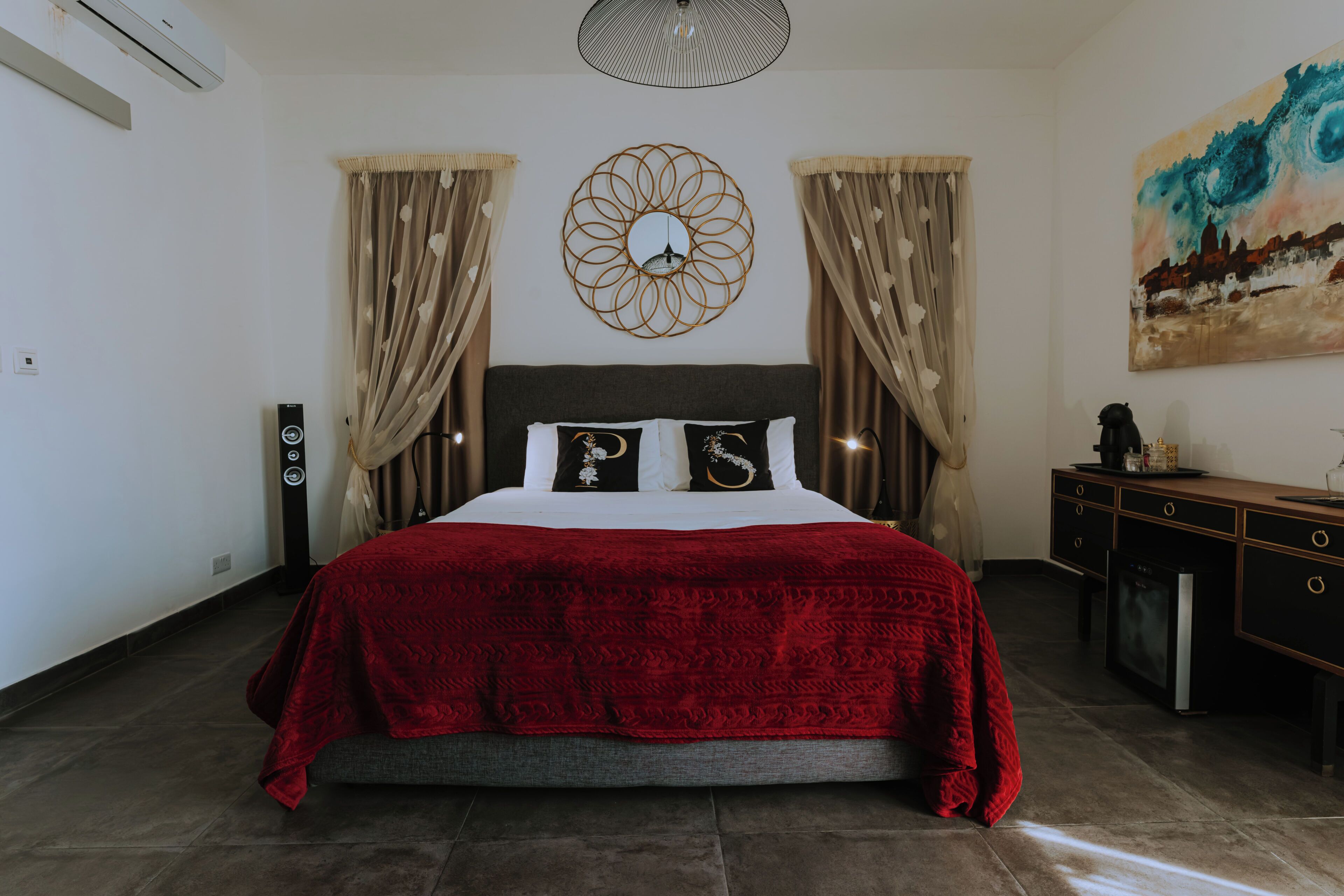 A bedroom with a bed, a wall-mounted mirror, a painting, and a desk with drawers.