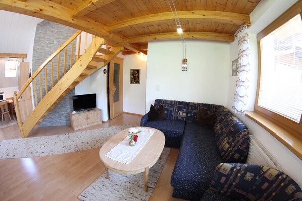 A cozy living room with a wooden staircase, a blue sofa, a wooden coffee table, and a television. - Gästehaus Burgi (Fuegen)