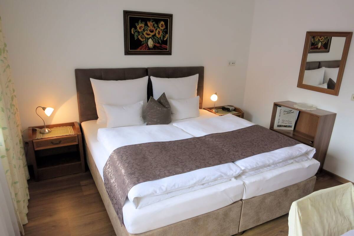 Cama bem arrumada com cabeceira marrom, roupa de cama branca e um travesseiro decorativo. Há duas mesas de cabeceira com abajures, um quadro emoldurado na parede e um espelho sobre a cama.
