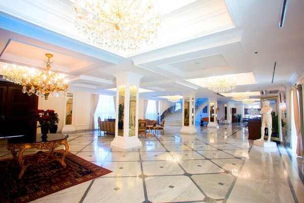 A spacious lobby with a marble floor, a grand chandelier, and a staircase leading to upper floors.