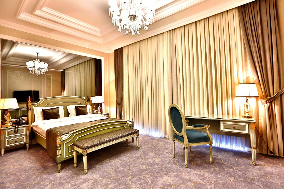 A hotel room with a large bed, a desk, a chair, a chandelier, and a large window with curtains.