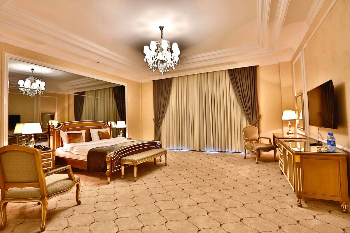 A hotel room with a large bed, a sitting area with a bench, a desk with a mirror, and a chandelier.