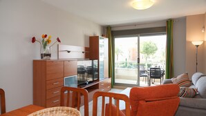 Ein modernes Wohnzimmer mit einer Couch, einem Esstisch mit Stühlen, einem Fernseher und einem Balkon mit Blick auf Gebäude und Bäume.