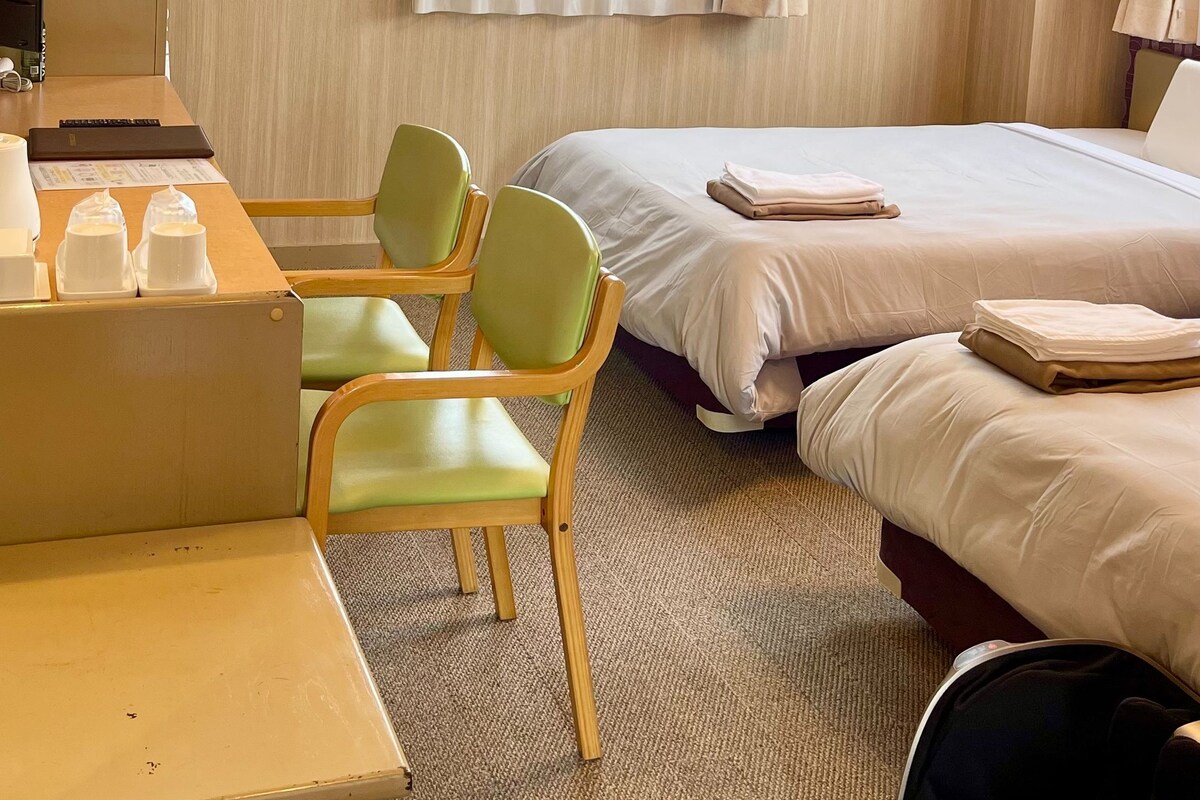 A hotel room with a bed, a desk with a chair, a small table with cups, a trash bin, and a window with curtains.