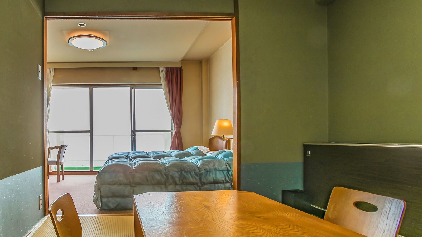 A hotel room with a bed, a wooden table, a chair, a lamp, and a view through a window.