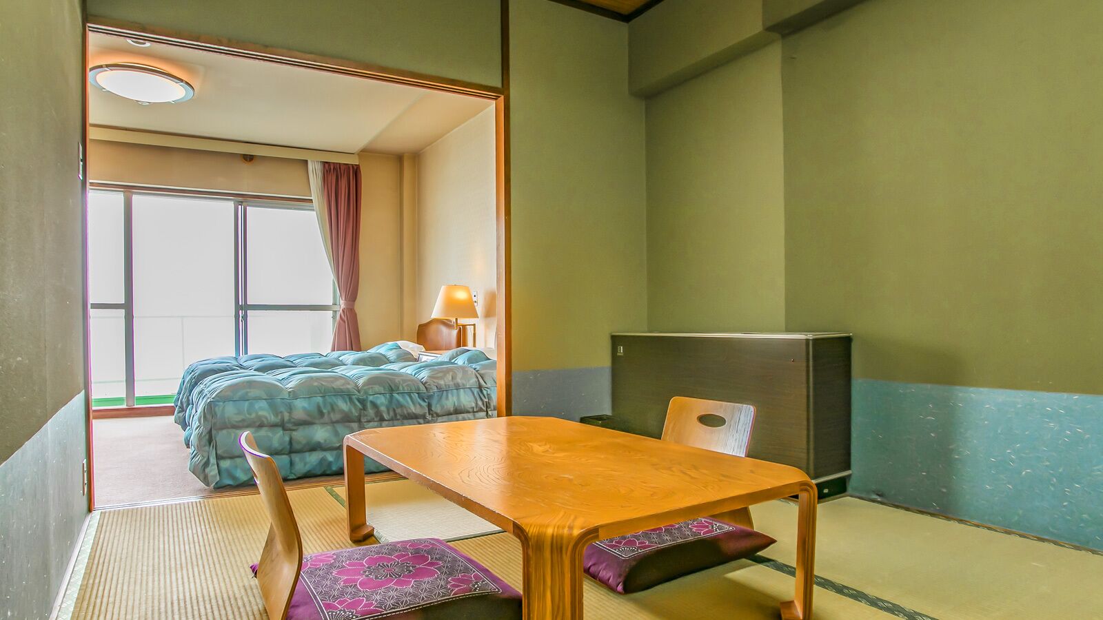 A room with a bed, a table, a chair, a lamp, and a refrigerator.