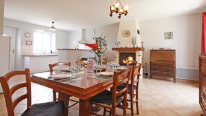 A dining area with a wooden table set for four, surrounded by chairs. A fireplace with a mantel is visible in the background, and a chandelier hangs above the table. - Family stay in the heart of Perche Park (La Chapelle-Souëf)