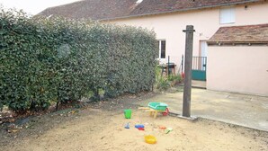 A residential backyard with a sandy play area, a green wheelbarrow, and a hedge. - Family stay in the heart of Perche Park (La Chapelle-Souëf)