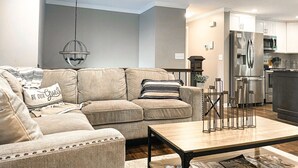 A living room with a beige sofa, a coffee table, and a kitchen area with a refrigerator and cabinets. - Fire Pit • Foosball • Trailside • Relaxing Covered Deck (Charlotte)