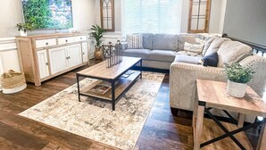 A living room with a sofa, coffee table, and TV, a kitchen with cabinets, and a dining area with a table and chairs. - Fire Pit • Foosball • Trailside • Relaxing Covered Deck (Charlotte)