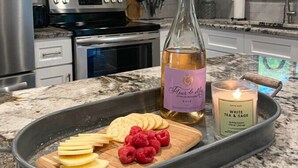 A kitchen with white cabinets, stainless steel appliances, a marble countertop, a bottle of Fleur de Lis wine, a candle, and a tray with cheese and raspberries. - Fire Pit • Foosball • Trailside • Relaxing Covered Deck (Charlotte)