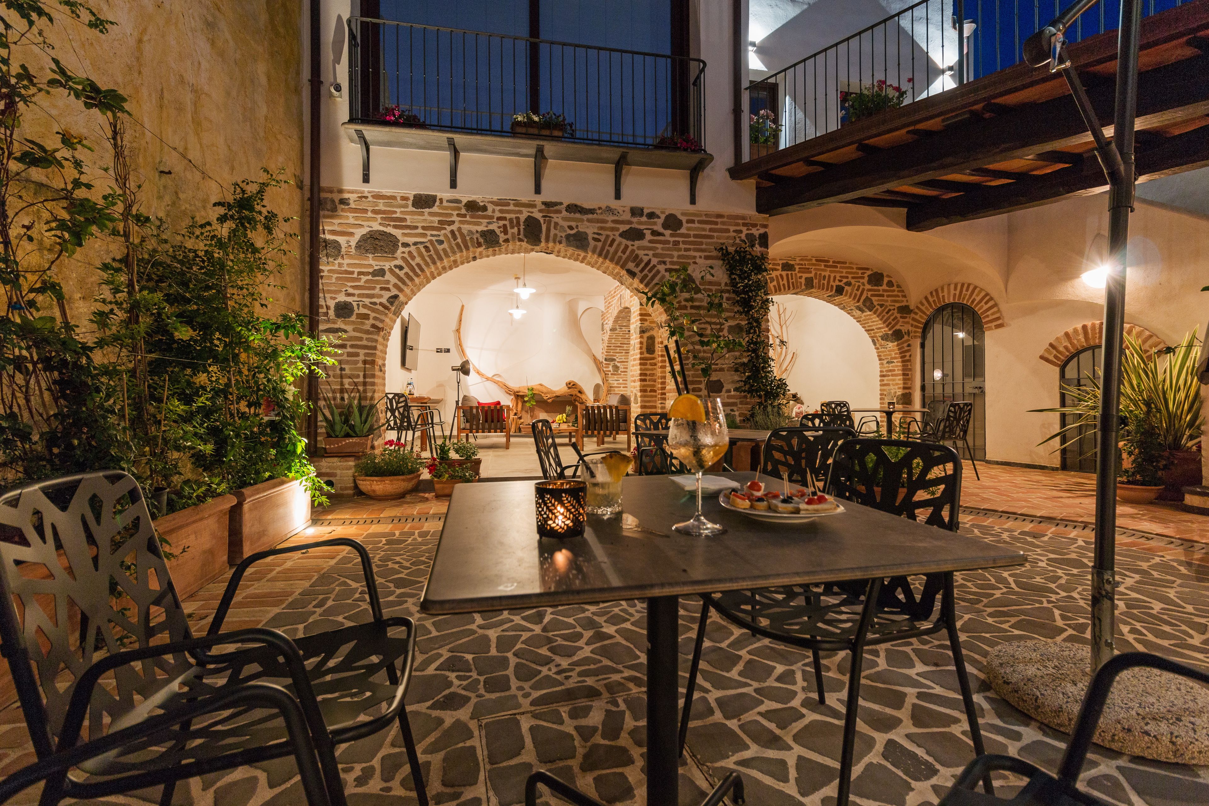 Une terrasse extérieure avec un sol en pierre, des tables et des chaises en métal, entourée de plantes en pot et d’un mur en pierre.