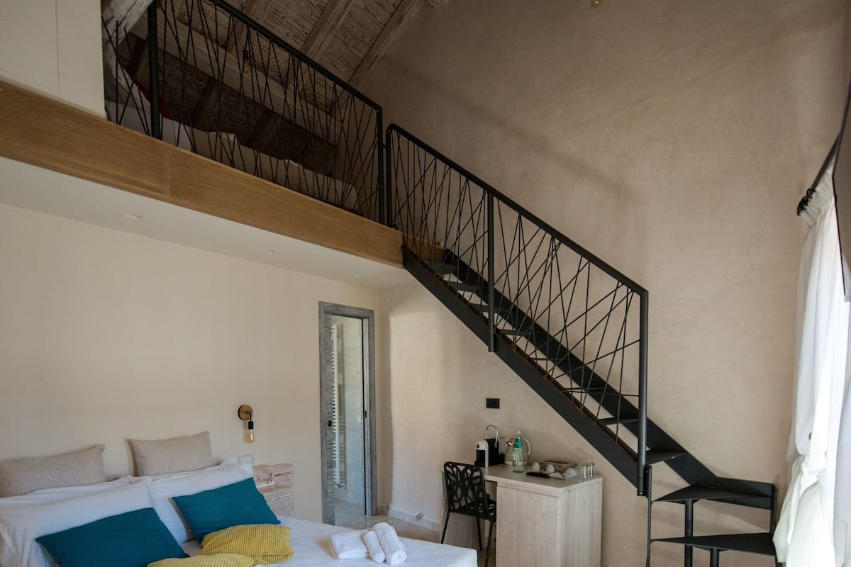 A bedroom with a large bed, a wooden ceiling, a staircase, and a small table with a chair.