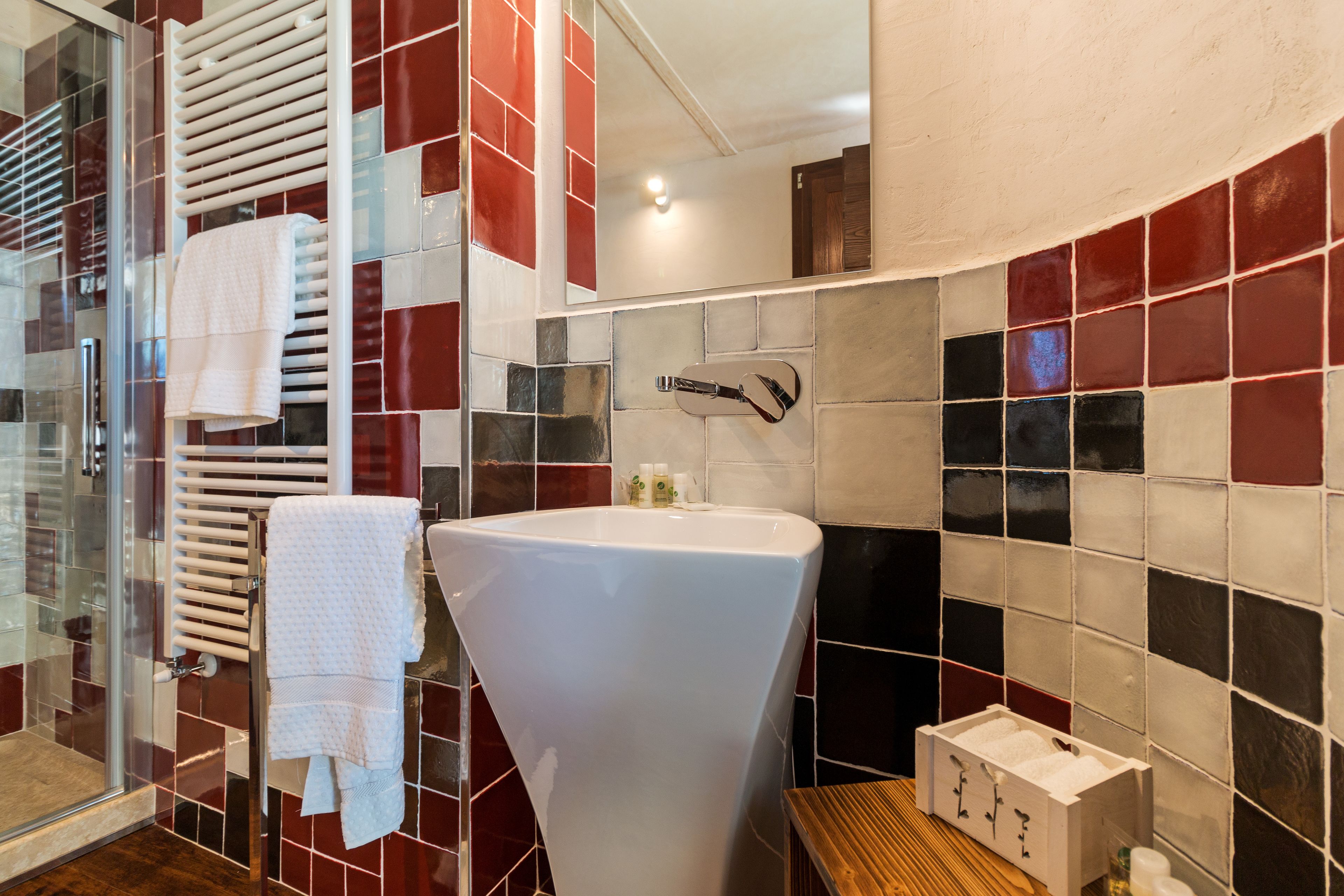 Een badkamer met een glazen douchecabine, een wit wastafeltje en een betegelde muur met rode en zwarte tegels.