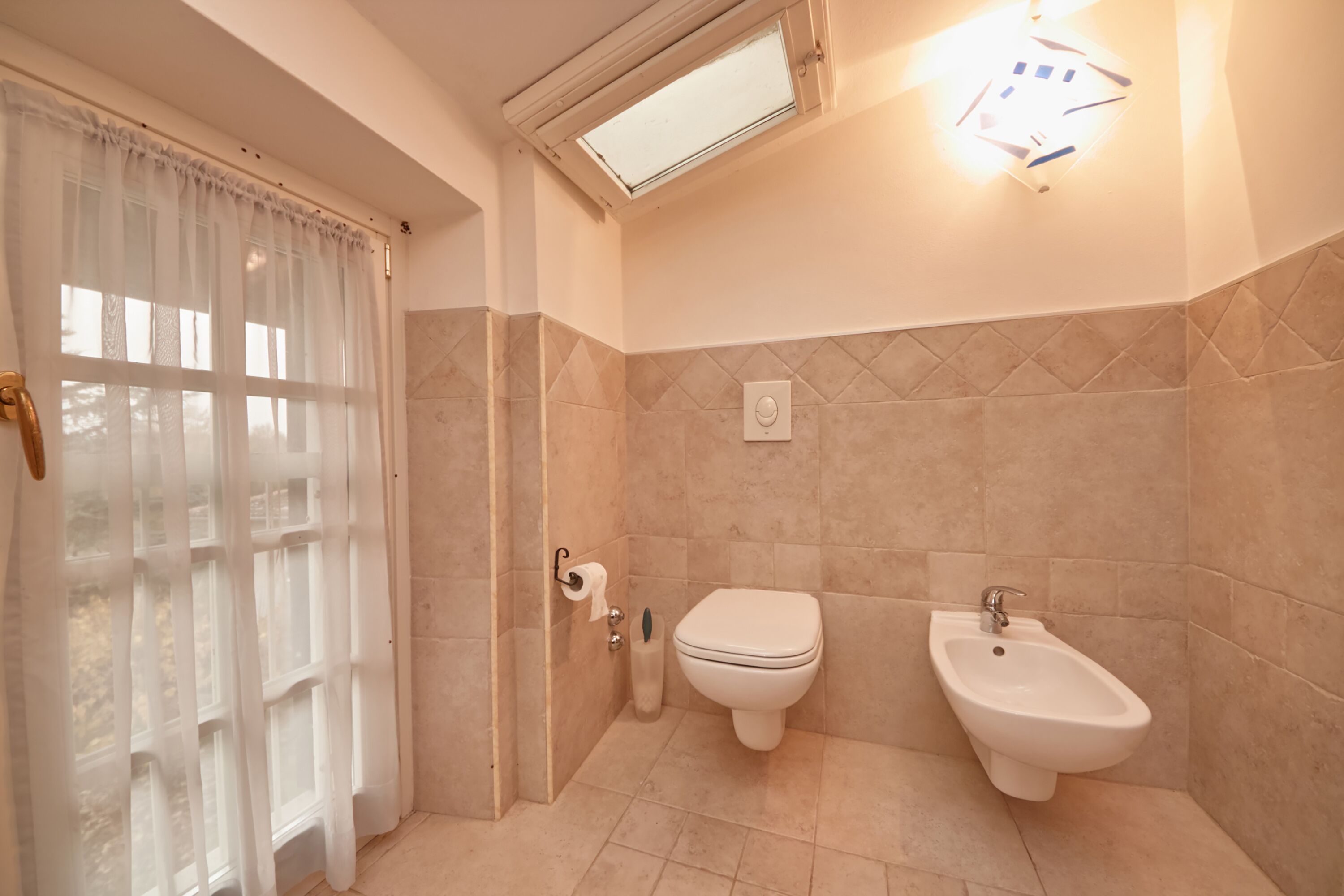 Et badeværelse med toilet, bidet og et vindue med lette gardiner.